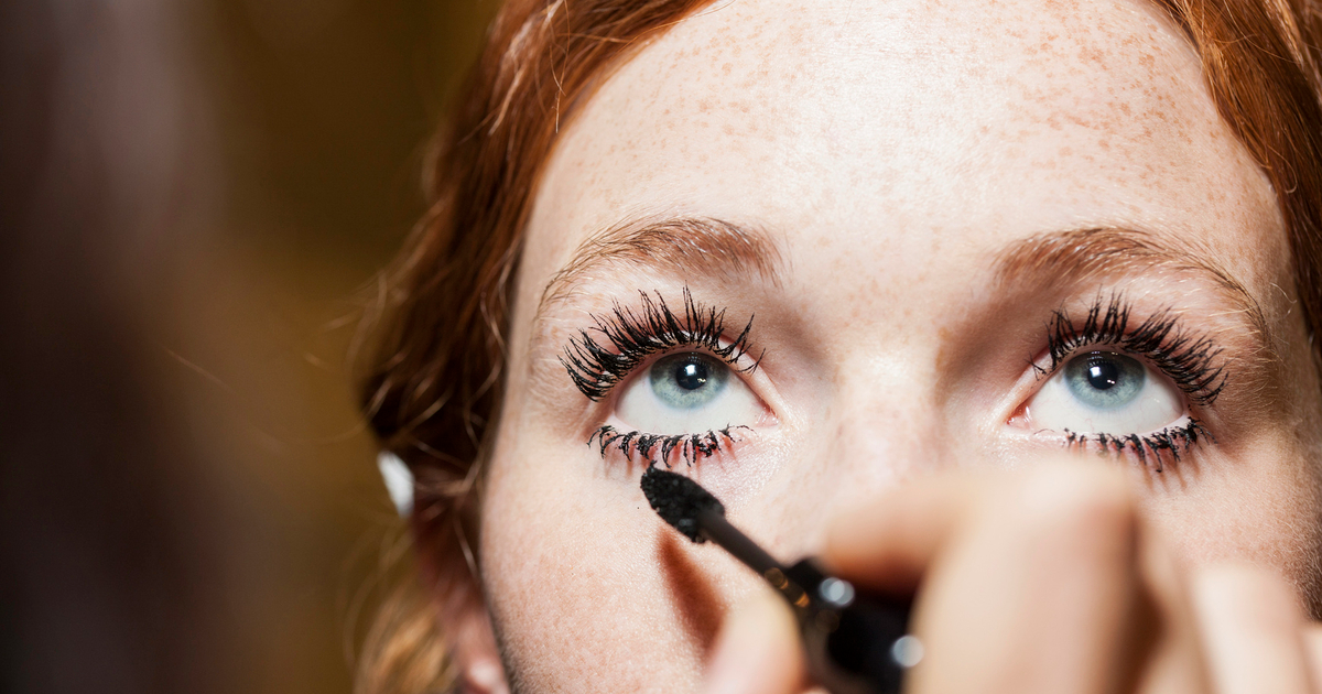 woman with red hair and eyebrows and blue eyes applying mascara to her long eyelashes