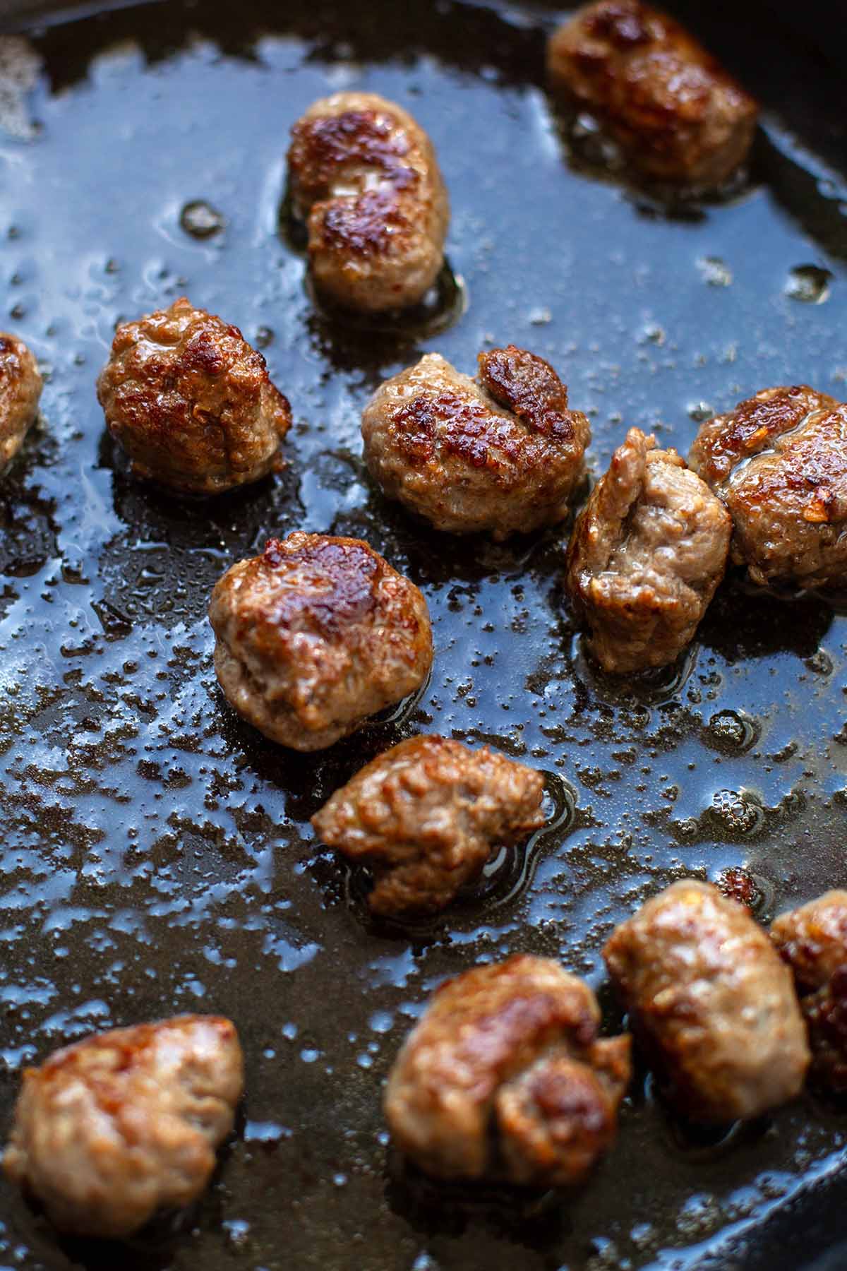 Coarse sausages rolled into balls are fried with oil in a black pan.