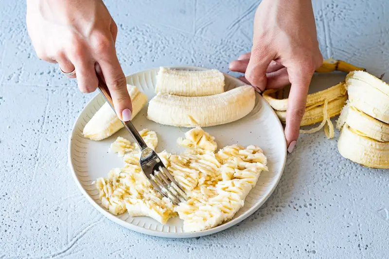 Mature bananas are crushed on a white plate with a fork.