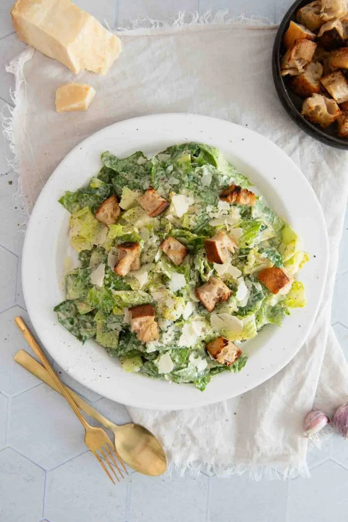 Classic Caesar Salad on a white plate: Roman salad in a creamy dressing, garnished with croutons and planed parmesan, next to it golden cutlery and a bowl with extra croutons.
